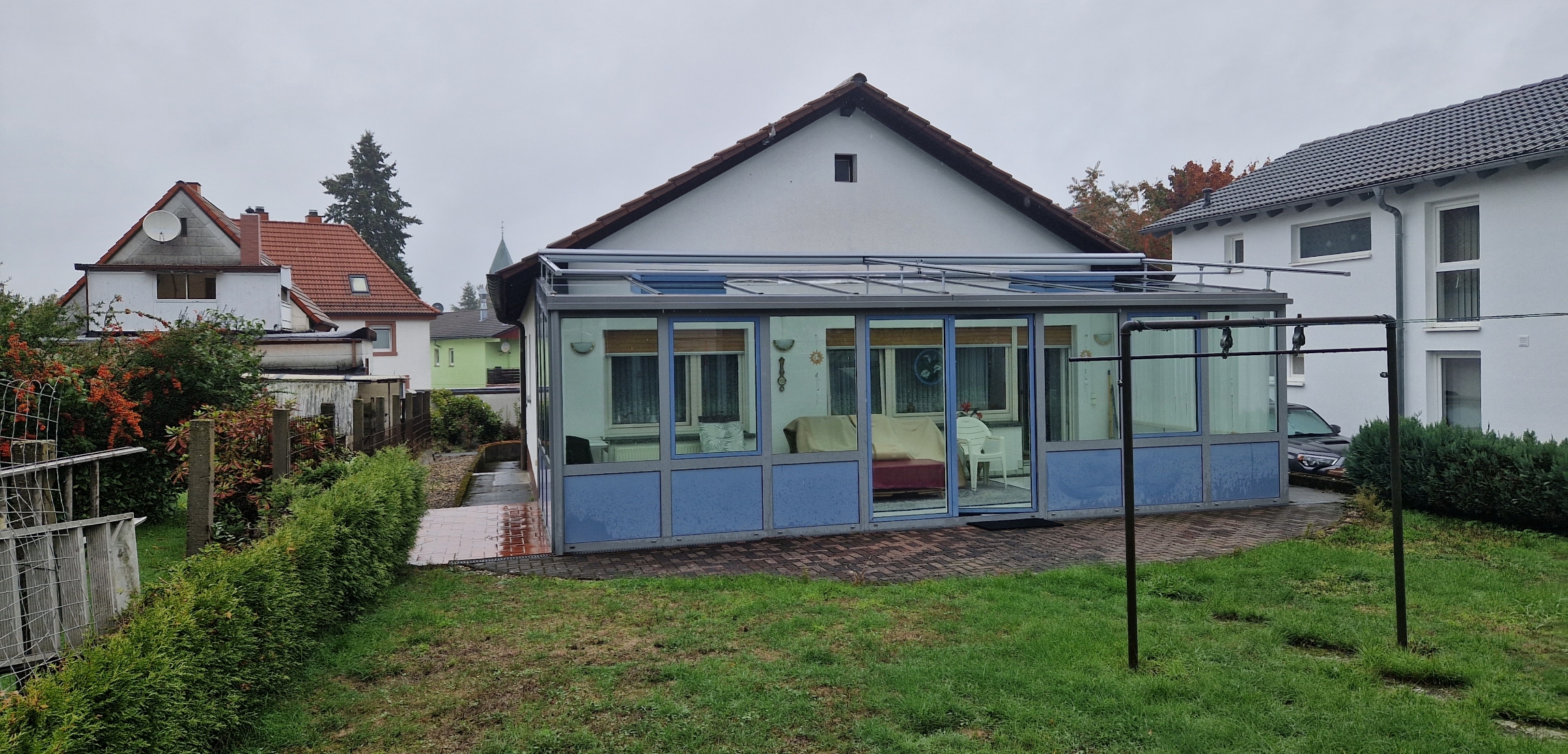 Vorderansicht eines weißen Hauses mit einer verglasten, blauen Terrasse und einem grünen Garten bei bewölktem Himmel.