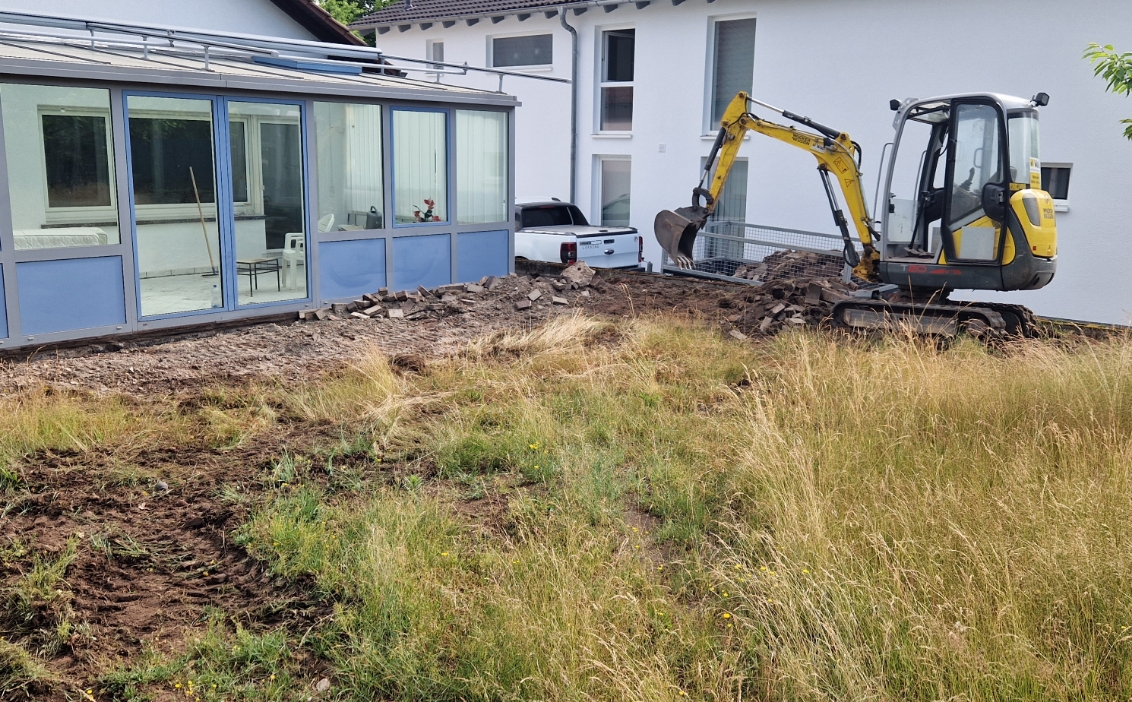 Gelber Minibagger gräbt Erde in einem Garten neben einem Haus mit verglaster Veranda und hohem Gras.
