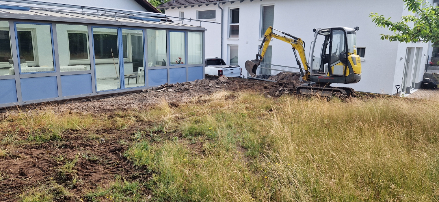 Bagger arbeitet auf einem unebenen Gartengrundstück neben einem weißen Gebäude mit Wintergarten und einem geparkten weißen Pick-up.