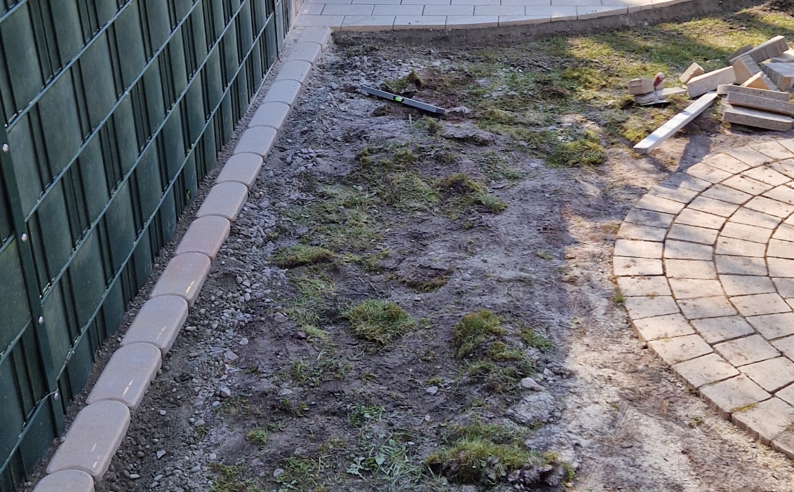 Ein teilweise gepflasterter Gartenweg mit einem halbkreisförmigen Steinsitzplatz und Boden mit Löchern und Grasflecken, daneben stapelweise Pflastersteine und eine Wasserwaage.