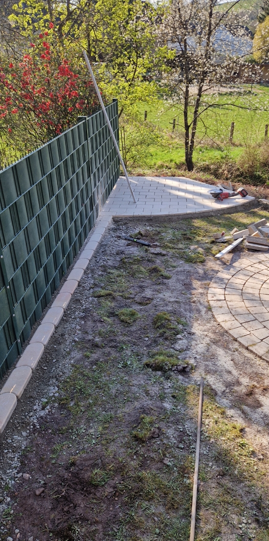 Im Garten sind neue Pflastersteine verlegt, ein Bereich ist noch nicht fertig, neben einem grünen Zaun und blühenden Bäumen im Hintergrund.