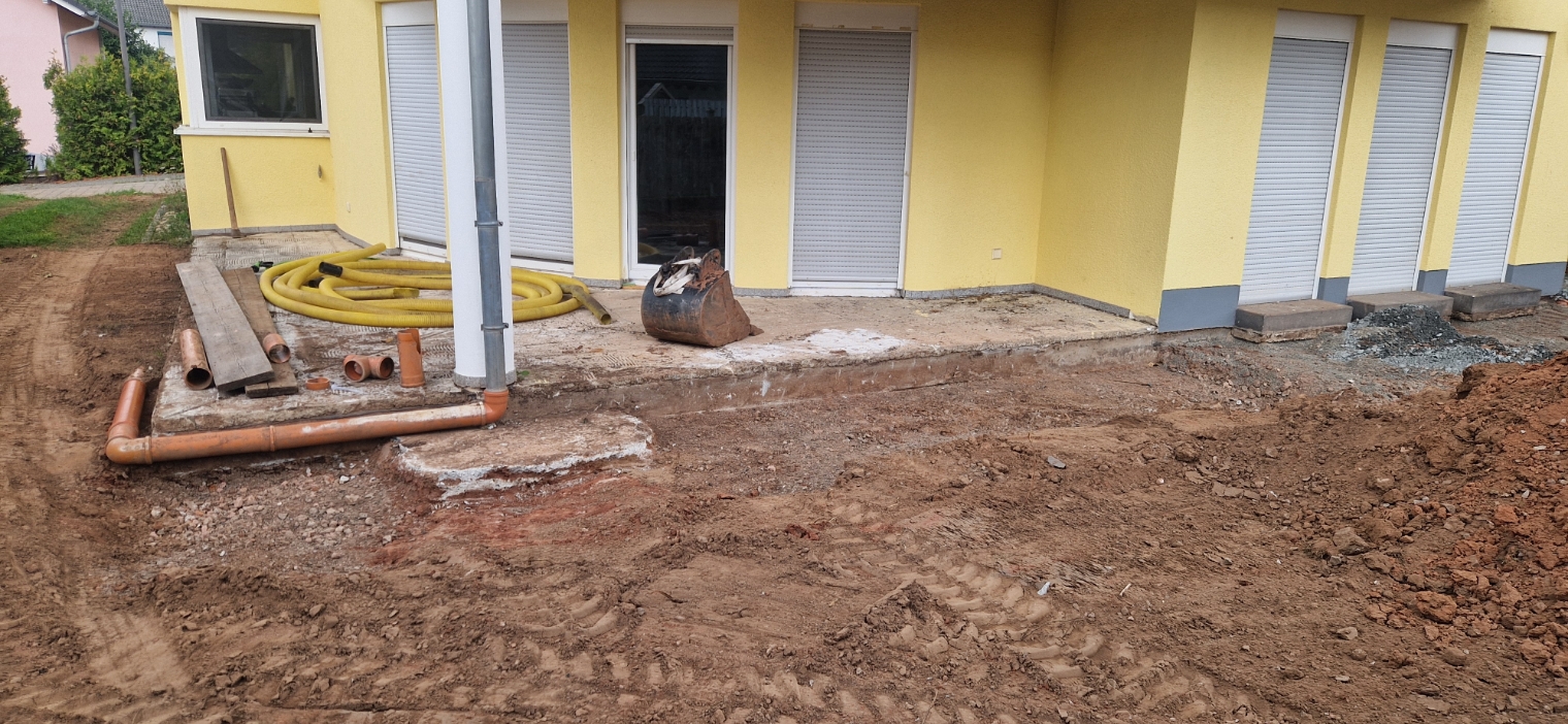 Haus mit gelber Fassade und weißen Rollläden, vor dem Eingang Baustelle mit Erde, Rohren, Holzbrettern und einem Baggerlöffel.