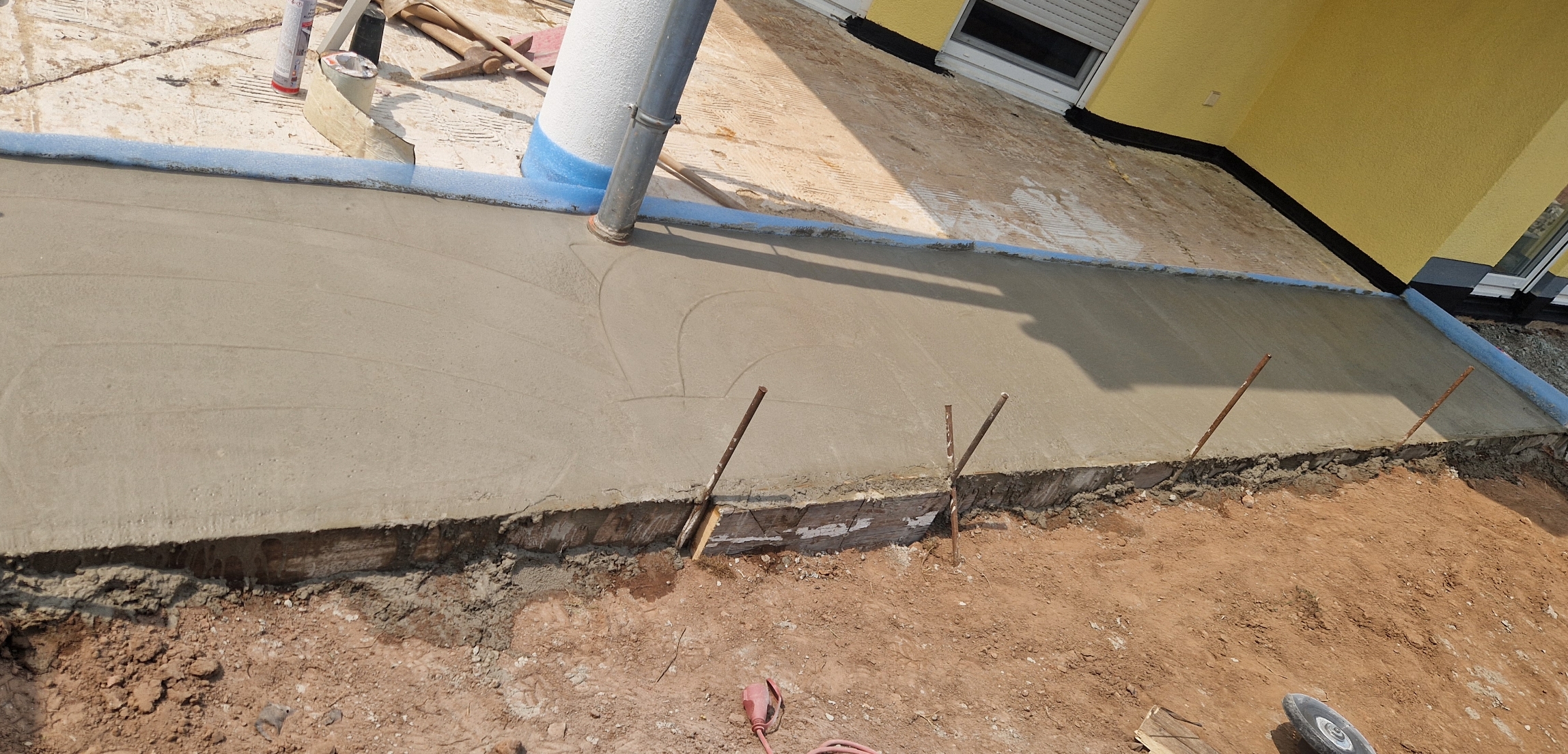 Frisch gegossener Beton für eine Terrasse vor einem gelben Haus mit Bauwerkzeugen im Hintergrund.