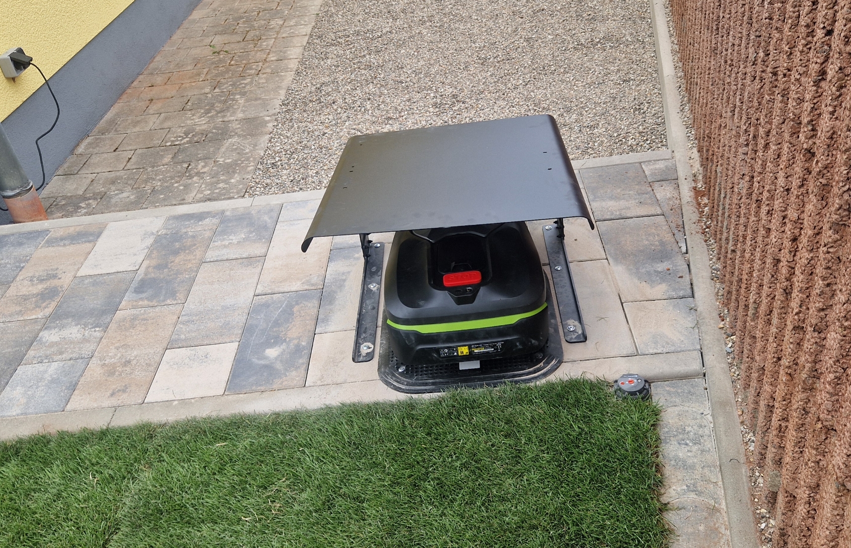 Roboter-Rasenmäher unter einem flachen Metallschutz auf einer gepflasterten Fläche neben einem Grasrasen.