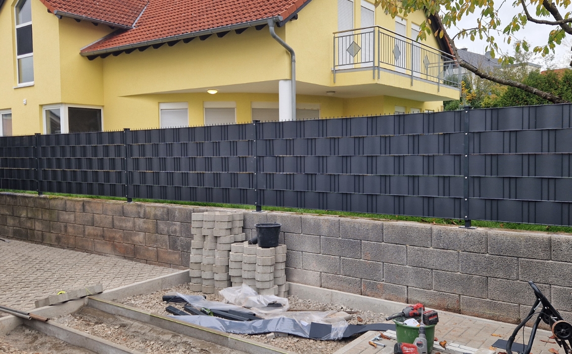 Eine Baustelle vor einem gelben Haus mit roten Dachziegeln, auf der graue Pflastersteine gestapelt sind und ein schwarzer Sichtschutzzaun auf einer Betonmauer installiert wurde.