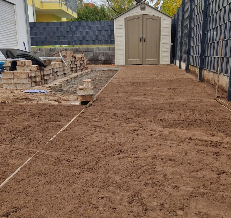 Gartenweg im Bau mit aufgestapelten Pflastersteinen und einem grauen Geräteschuppen am Ende.