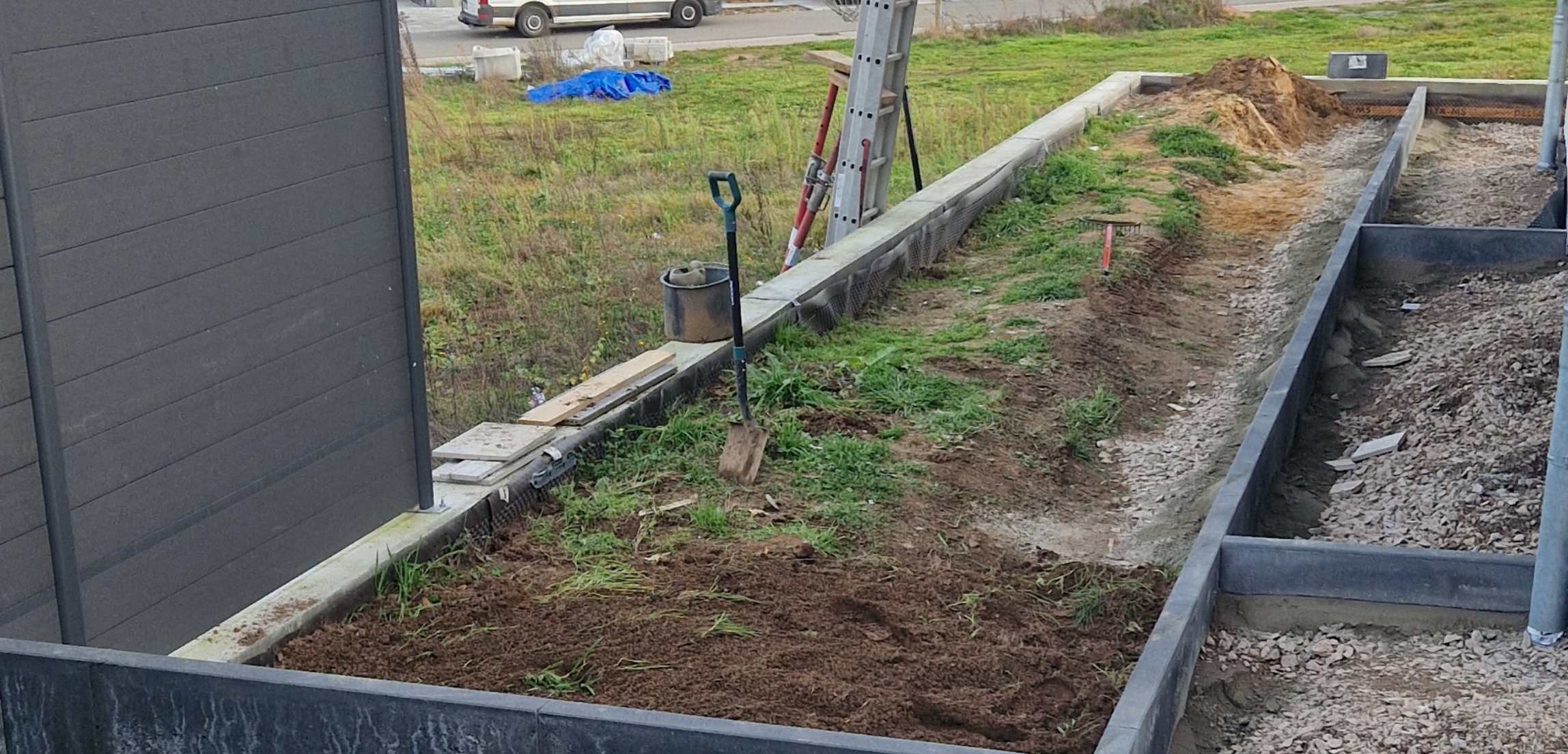 Baustelle mit Erdarbeiten, einem blauen Gartenschlauch, einer Schaufel und einem silbernen Transportwagen im Hintergrund.