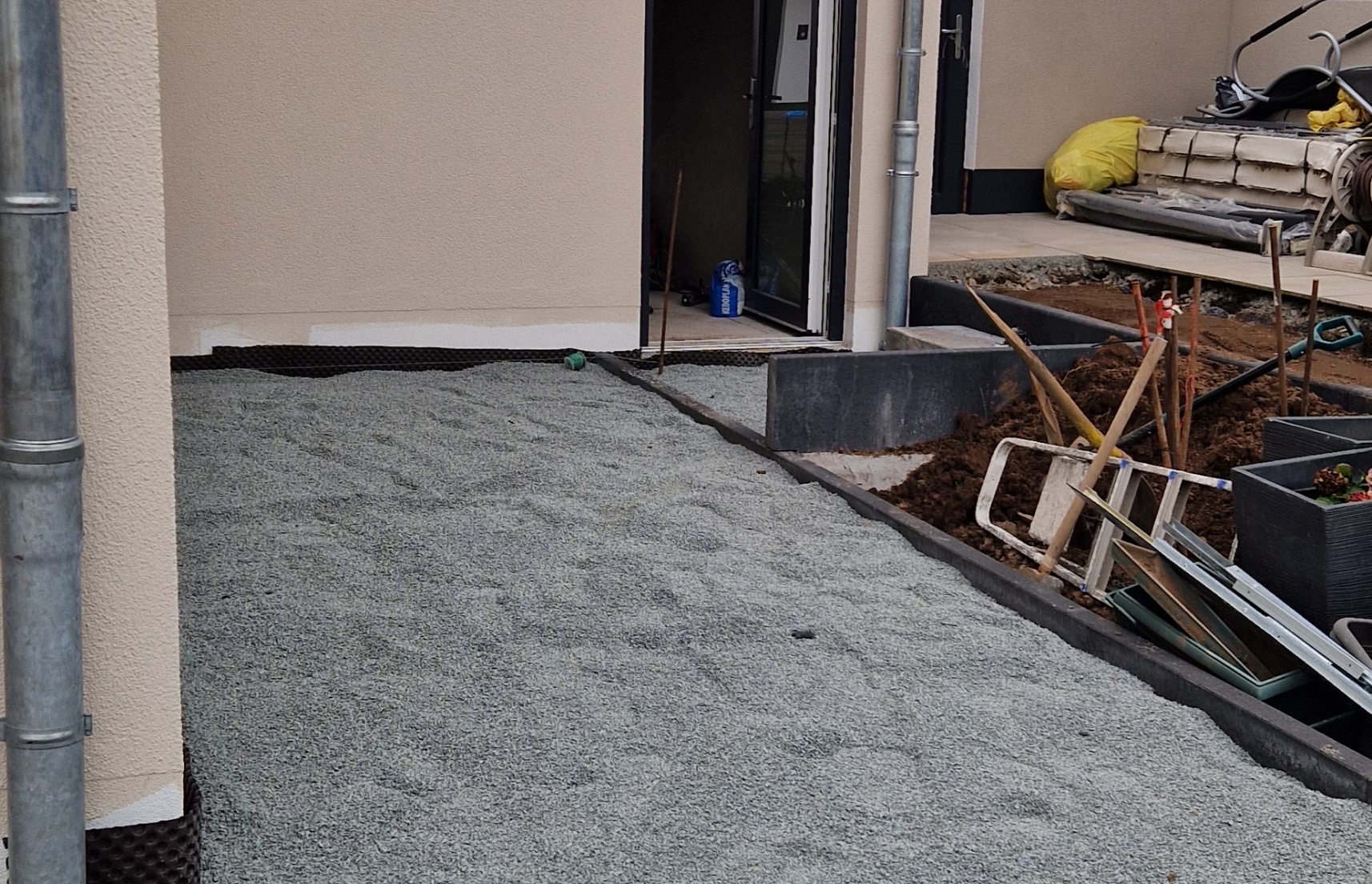 Bauarbeiten mit einer geschotterten Fläche neben einem Haus und Gartengeräten neben einem Hochbeet mit Erde.