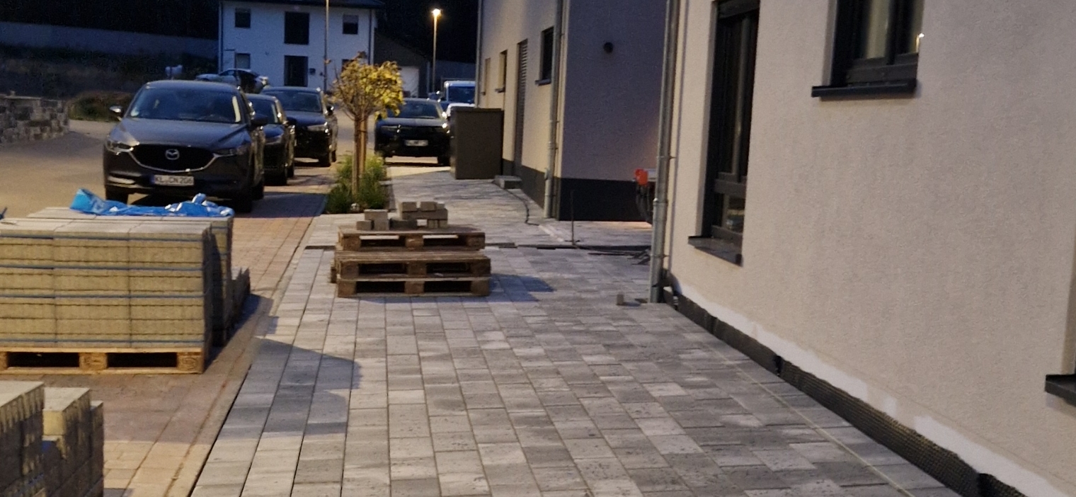 Gepflasterter Gehweg neben einem Haus mit gestapelten Paletten und Baustoffen, parkenden Autos und einer kleinen Baumreihe bei Dämmerung.
