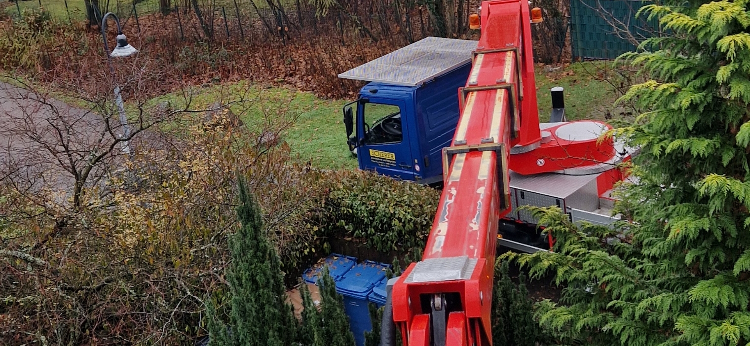 Roter Kranarm eines Lastwagens, der über eine Gartenlandschaft mit Bäumen und zwei blauen Mülltonnen hinausragt.
