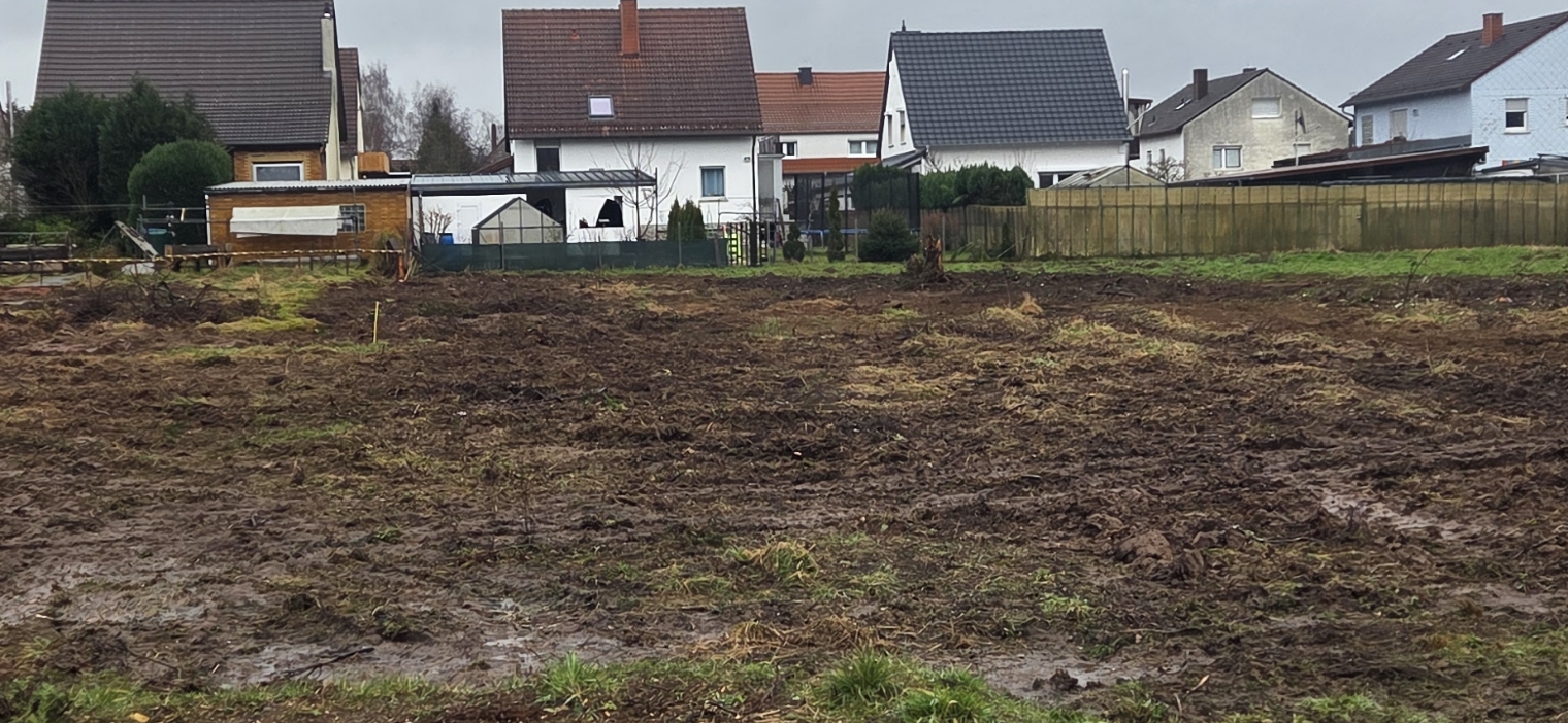 Ein nasses, unbebautes Feld mit rutschigem Boden vor einer Reihe von Wohnhäusern unter bewölktem Himmel.