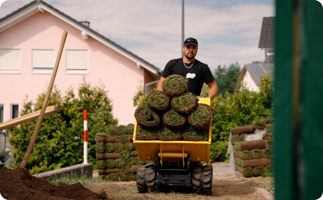 Mann schiebt eine gelbe Schubkarre mit aufgerolltem Rollrasen vor einem Haus im Garten.