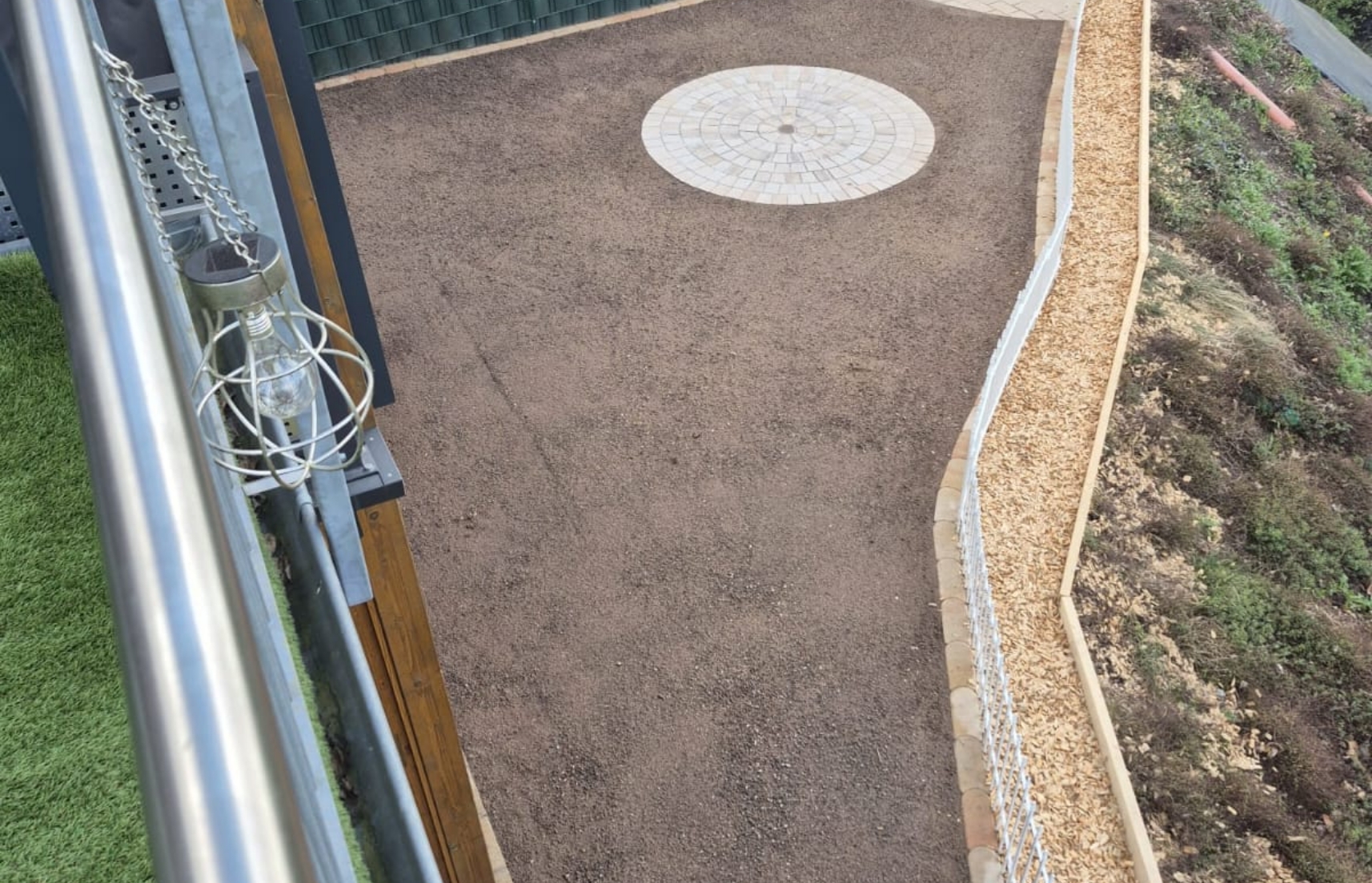 Blick von einem Balkon auf einen mit braunem Kies belegten Hof, einem runden Pflastermuster in der Mitte, einem Holzspalthaufen rechts und einem Metallgeländer links.